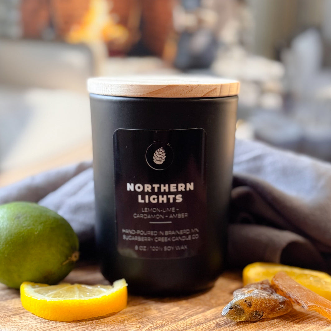 Black candle labeled 'Northern Lights' on a wooden board with fruit, in front of a fireplace.