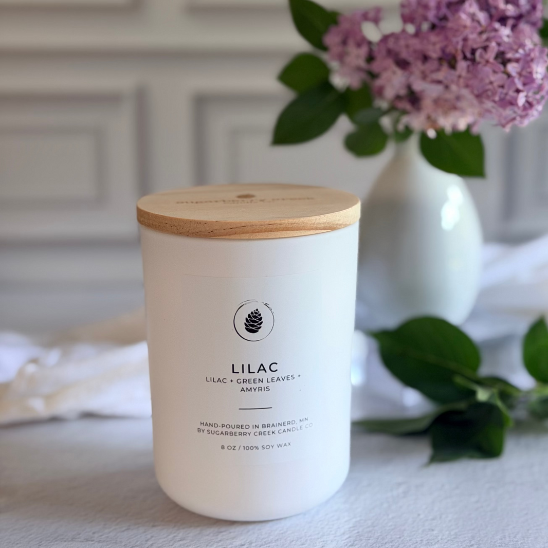 Candle labeled 'Lilac' with a wooden lid on a surface, next to a vase with purple flowers.