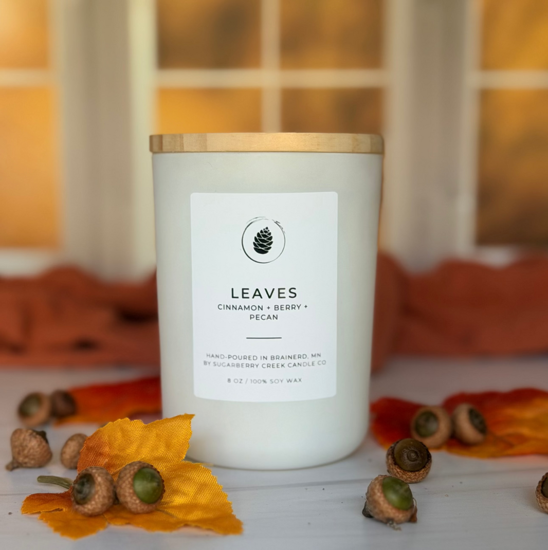 A white soy wax candle with a gold lid and a white label reading 'LEAVES Cinnamon + Berry + Pecan' sits on a white surface with scattered acorns and autumn leaves, against a warm, blurred window background.