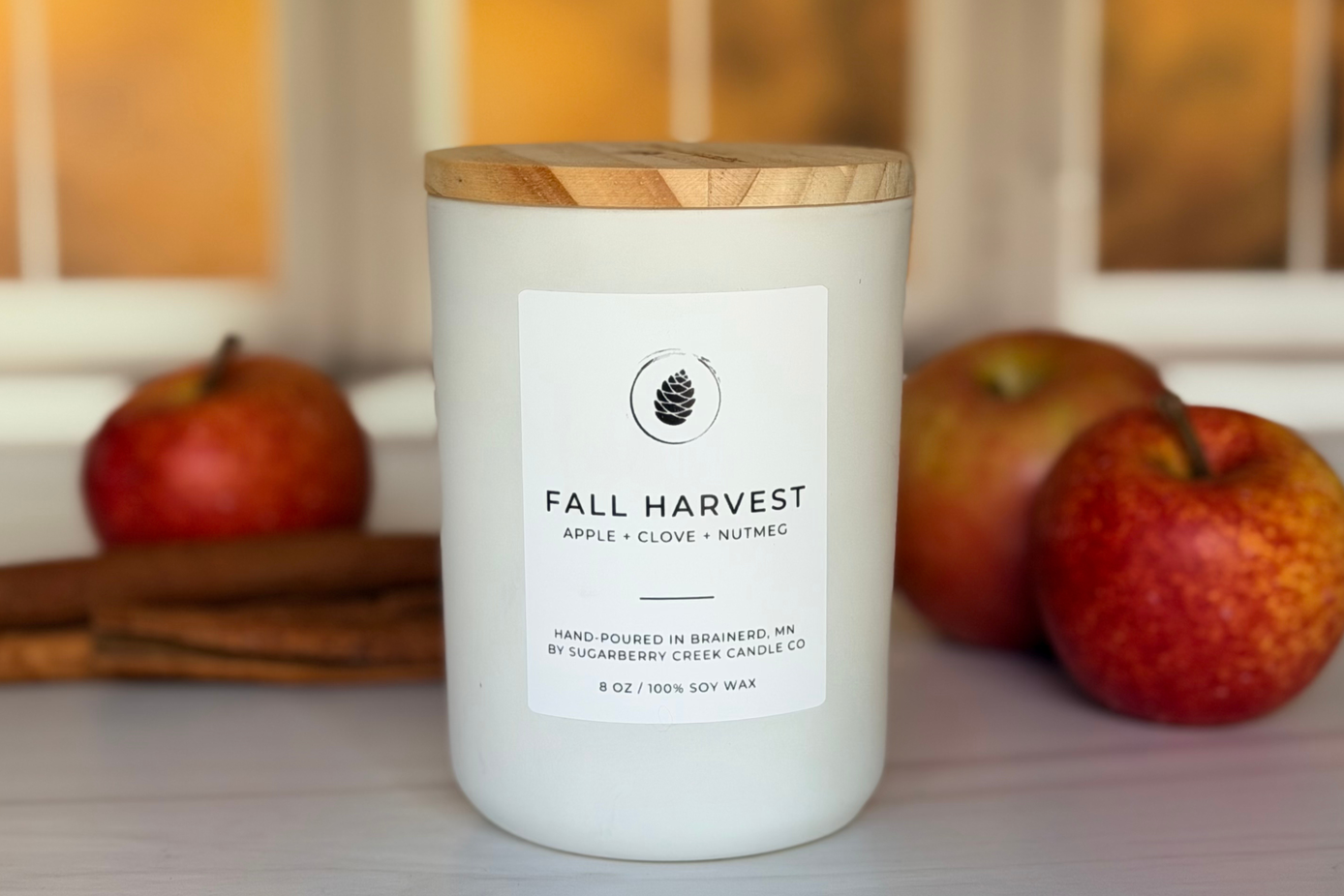 A white soy wax candle jar with a light wood lid, labeled 'FALL HARVEST' by Branded in Brimley, set on a light surface with red apples and cinnamon sticks in the background.