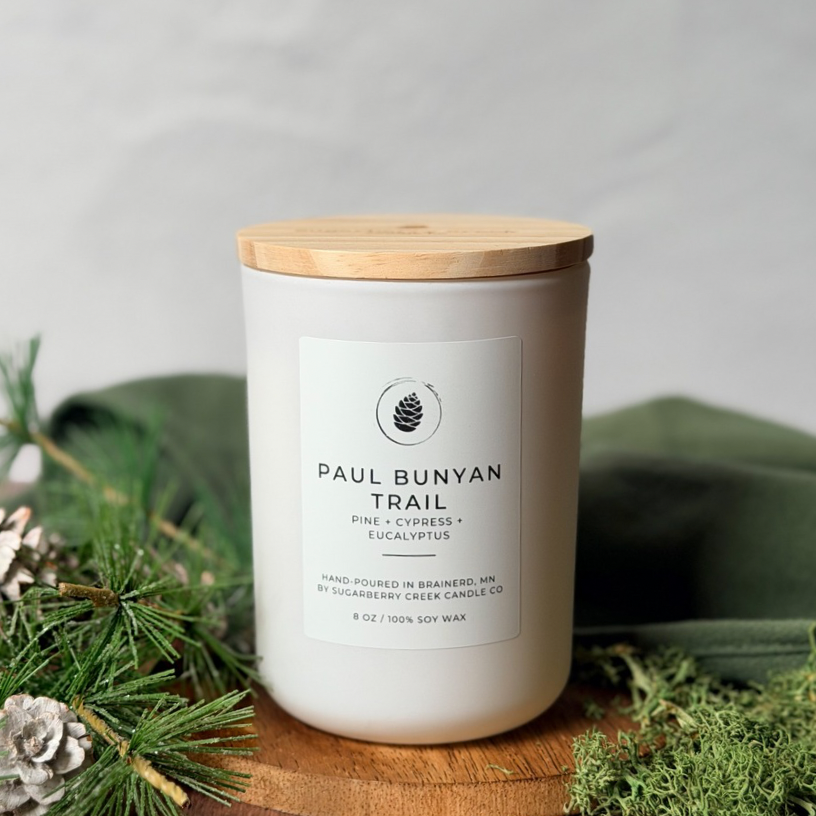 White candle with wooden lid on a wooden coaster surrounded by greenery