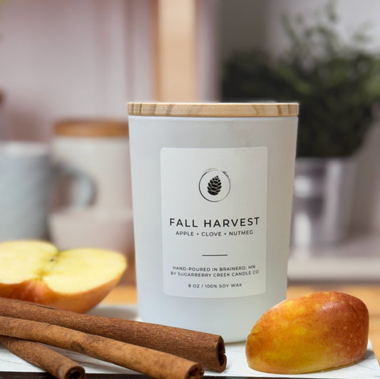 Candle labeled 'Fall Harvest' with apples, cinnamon sticks, and a loaf of bread on a table.