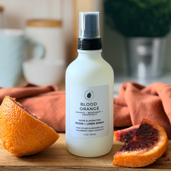 White spray bottle labeled 'Blood Orange' on a wooden cutting board with blood oranges, in a kitchen setting.