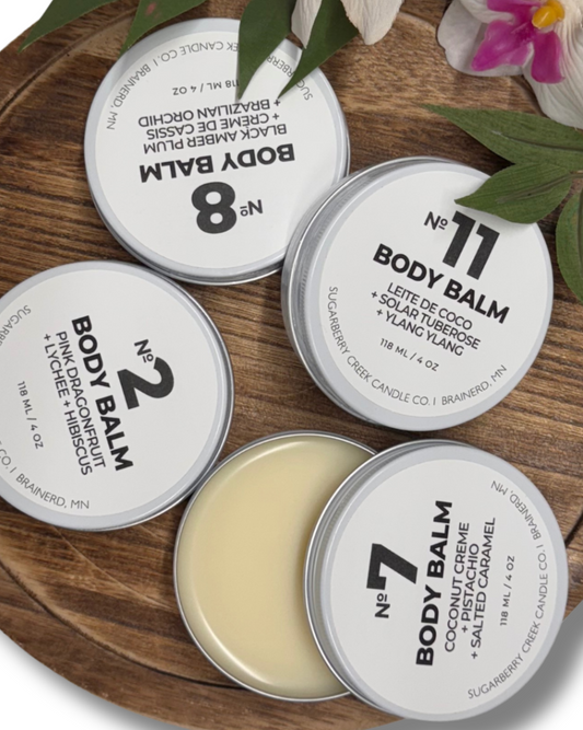 Collection of body balms in tins on a wooden surface with green leaves and flowers.