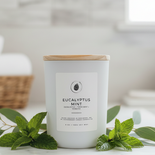 Eucalyptus Mint candle with wooden lid on a white surface with green leaves.