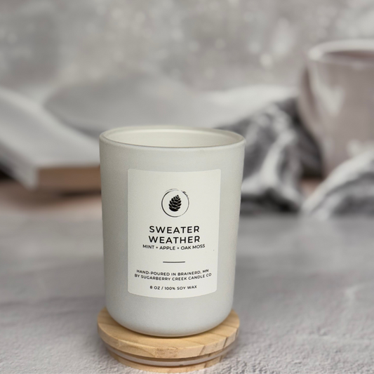 White candle sitting on a wood cover with a book and coffee cup in the background