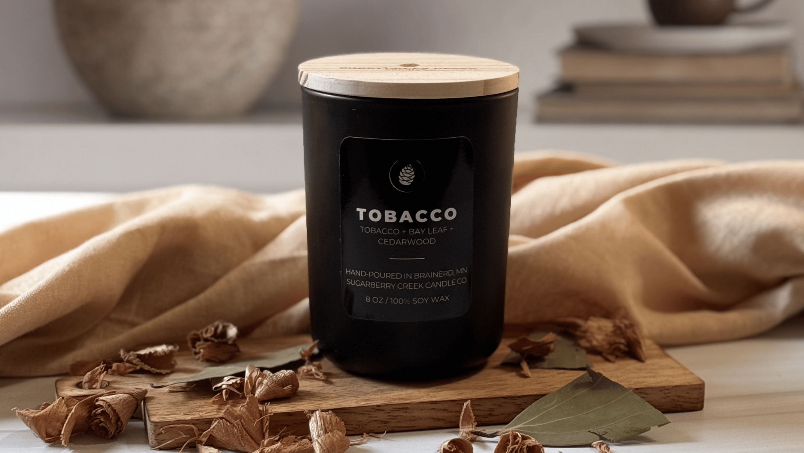 A black glass candle jar with a light wood lid sits on a wooden board surrounded by dried botanicals and a beige cloth, with a softly blurred indoor background.