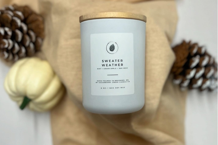 white candle jar on a tan towel surrounded by awhite pumpkin and pine cones