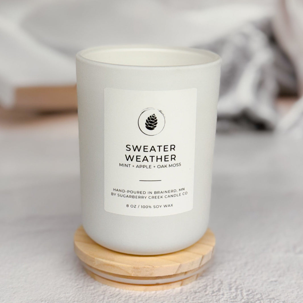 A white candle jar labeled 'SWEATER WEATHER' by The Pine Cone Candle Co., resting on a small round wooden coaster against a soft, blurred light gray background with a book and mug in the distance.