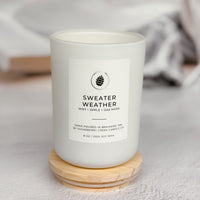 A white candle jar labeled 'SWEATER WEATHER' by The Pine Cone Candle Co., resting on a small round wooden coaster against a soft, blurred light gray background with a book and mug in the distance.