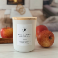 A white jar candle with a light wood lid labeled 'FALL HARVEST' sits on a tabletop in a cozy indoor setting, with apples and cinnamon sticks around it and a blurred living room background.
