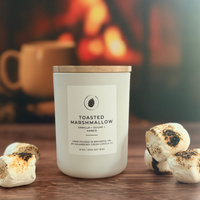 A frosted glass candle jar labeled 'TOASTED MARSHMALLOW' sits on a wooden surface with toasted marshmallows placed around it. The background shows a warm, blurred fireplace scene with a mug, creating a cozy indoor atmosphere.