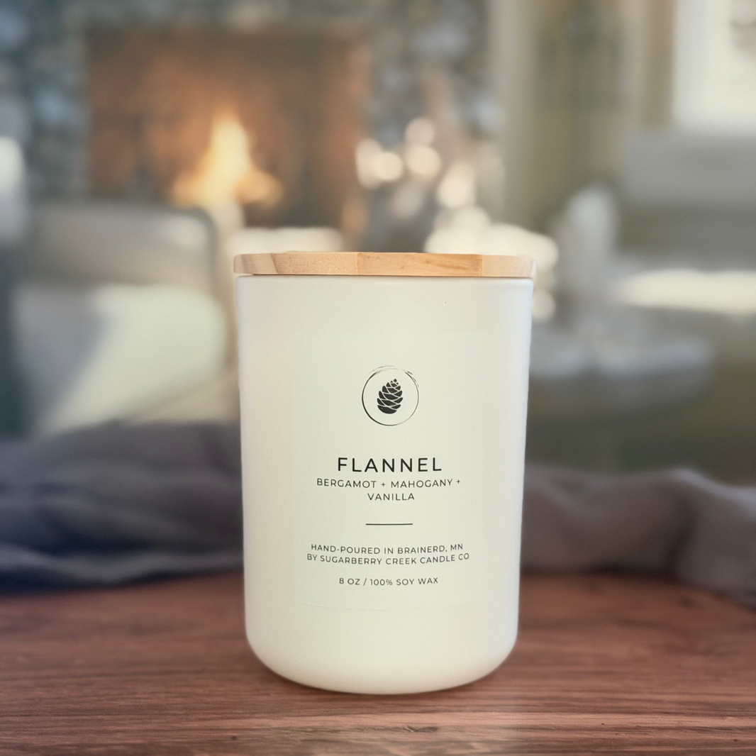A white soy wax candle jar with a light wood lid sits on a wooden table in a cozy, blurred living room setting with a fireplace in the background. The label reads 'FLANNEL' and 'BENJAMIN + MANGOLDY + VANILLA'.