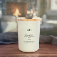 A white soy wax candle jar with a light wood lid sits on a wooden table in a cozy, blurred living room setting with a fireplace in the background. The label reads 'FLANNEL' and 'BENJAMIN + MANGOLDY + VANILLA'.