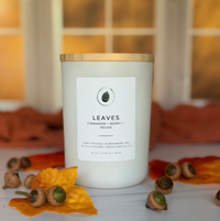 A white soy wax candle with a gold lid and a white label reading 'LEAVES Cinnamon + Berry + Pecan' sits on a white surface with scattered acorns and autumn leaves, against a warm, blurred window background.