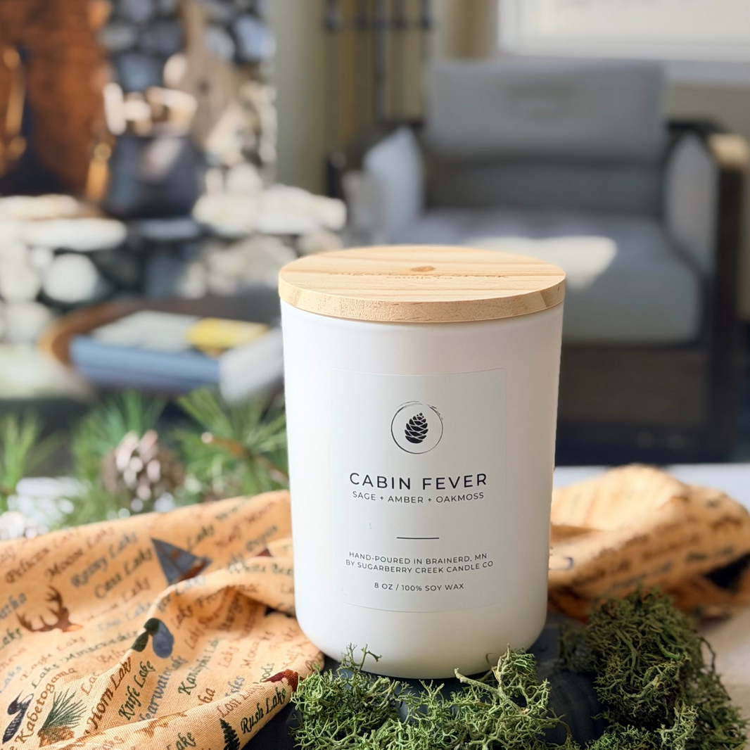 Candle labeled 'Cabin Fever' on a table with a cozy indoor setting featuring a fireplace.