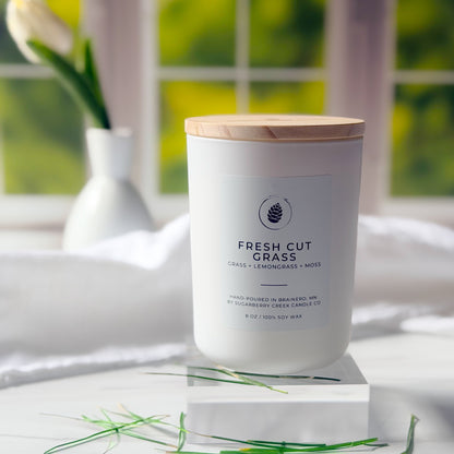 Candle labeled 'Fresh Cut Grass' on a white surface with a blurred green outdoor background