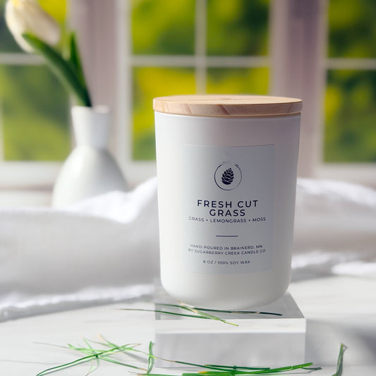 Candle labeled 'Fresh Cut Grass' on a white surface with a blurred green outdoor background