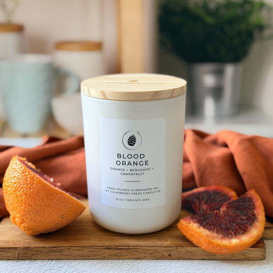 Candle labeled 'Blood Orange' on a wooden board with sliced oranges, set against a blurred indoor background.