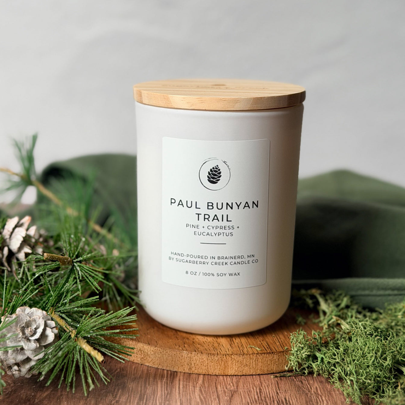 Candle labeled 'Paul Bunyan Trail' with a wooden lid on a wooden surface with greenery.