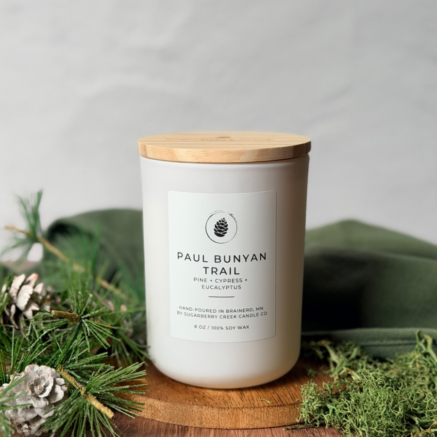 White candle with wooden lid on a wooden coaster surrounded by greenery