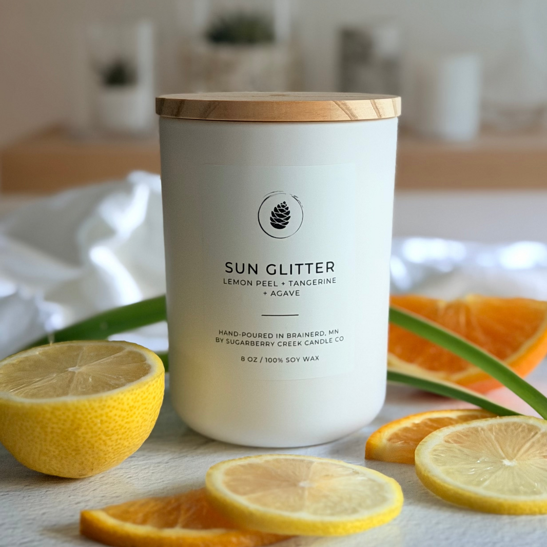 Candle labeled 'Sun Glitter' with lemon slices and an orange on a white surface.