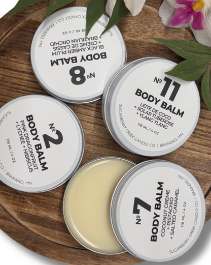 Collection of body balms in tins on a wooden surface with green leaves and flowers.