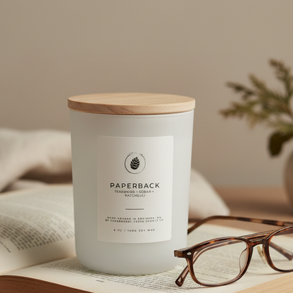 Candle labeled 'Paperback' on an open book with glasses and a plant in the background
