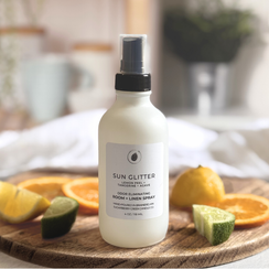 Bottle of Sun Glitter room and linen spray on a wooden board with citrus fruits.
