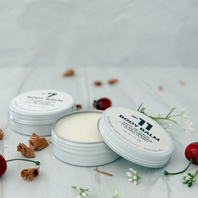 No. 11 white body balm tin, with the cover off, in front with a No. 7 white body balm tin behind it, surrounded by white flowers, cherriess and wood shavings